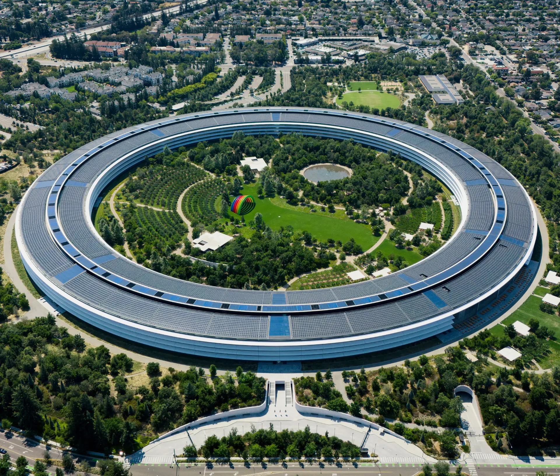 Aerial view of a large, circular, futuristic office building surrounded by greenery, trees, and pathways, with a cityscape in the background. The scene highlights productivity as remote teams collaborate seamlessly inside the innovative structure.