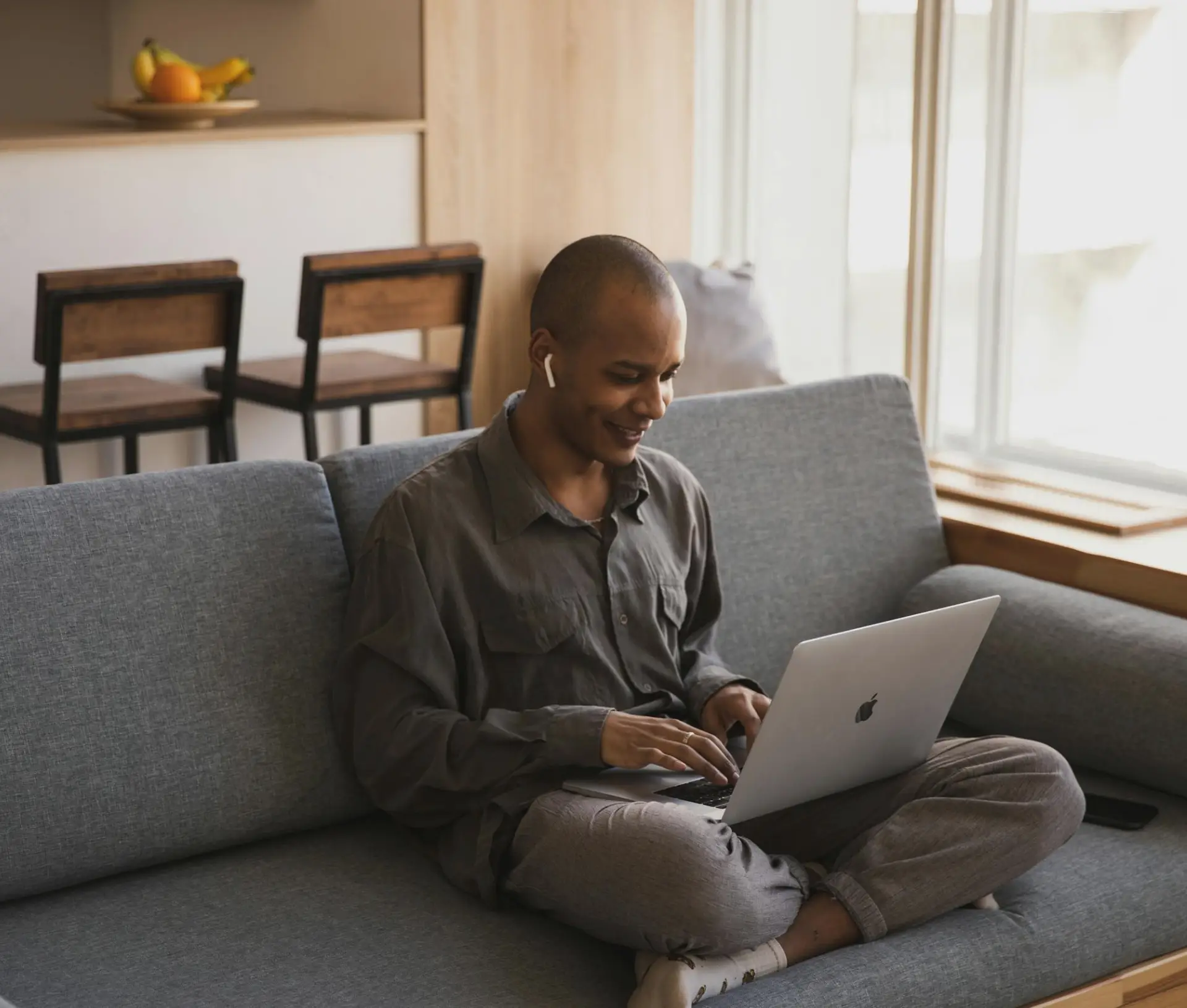 A person sits cross legged on a grey sofa, smiling while using a laptop running macOS. They are wearing casual clothes and wireless earphones. Sunlight streams in from a large window, and a fruit bowl is on a shelf in the background.