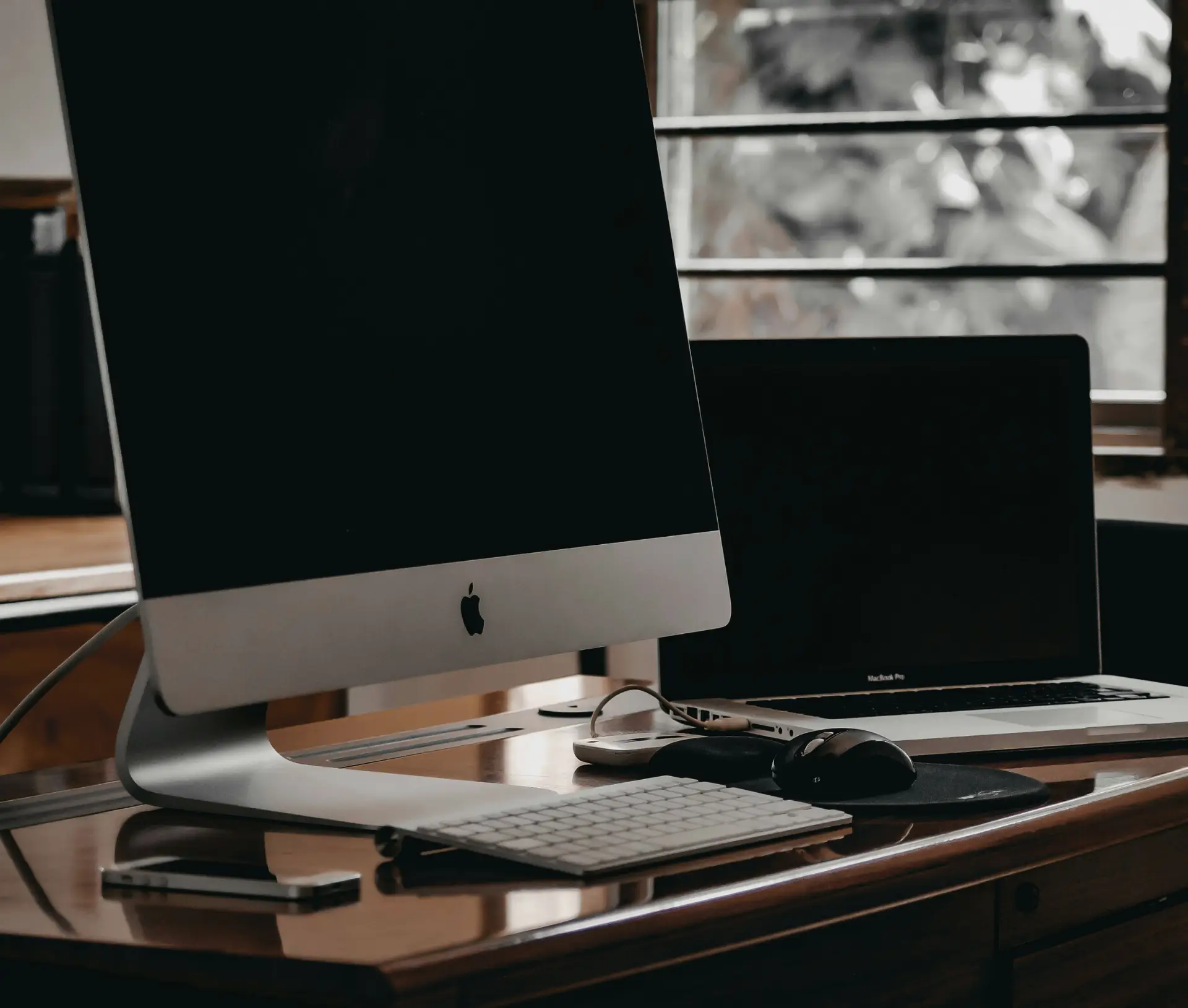 A desktop iMac, wireless keyboard, mouse, spectacles, and a closed MacBook are arranged on a wooden desk near a window with a blurred outdoor view, ideal for managing Apple Business MDM tasks with Jamf or Addigy.