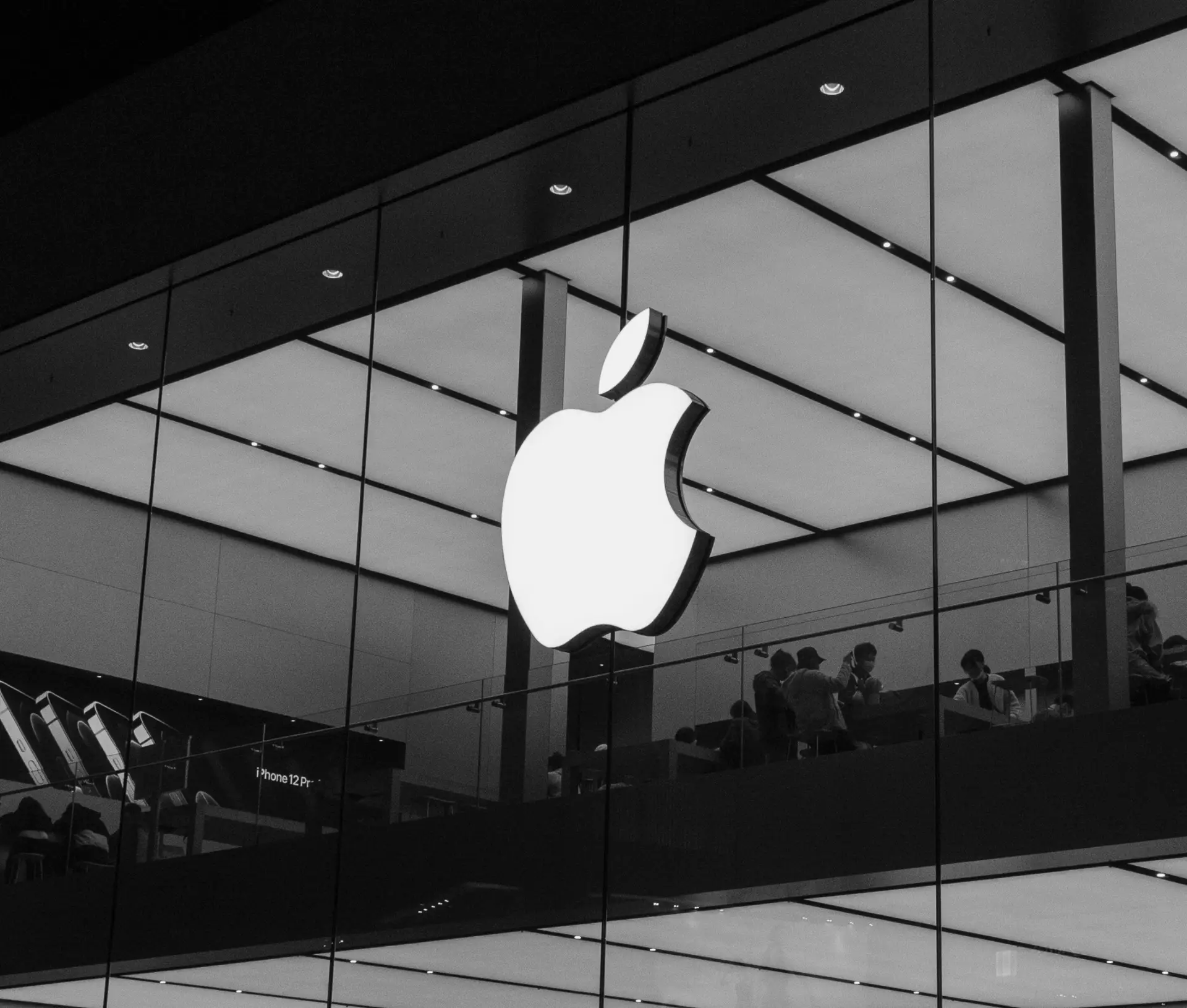 A large, illuminated Apple logo on the glass façade of an Apple Store, showcasing people inside the modern, brightly lit interior—an inspiring space where organisation and innovation define the all in one platform for success. The photo is in black and white.