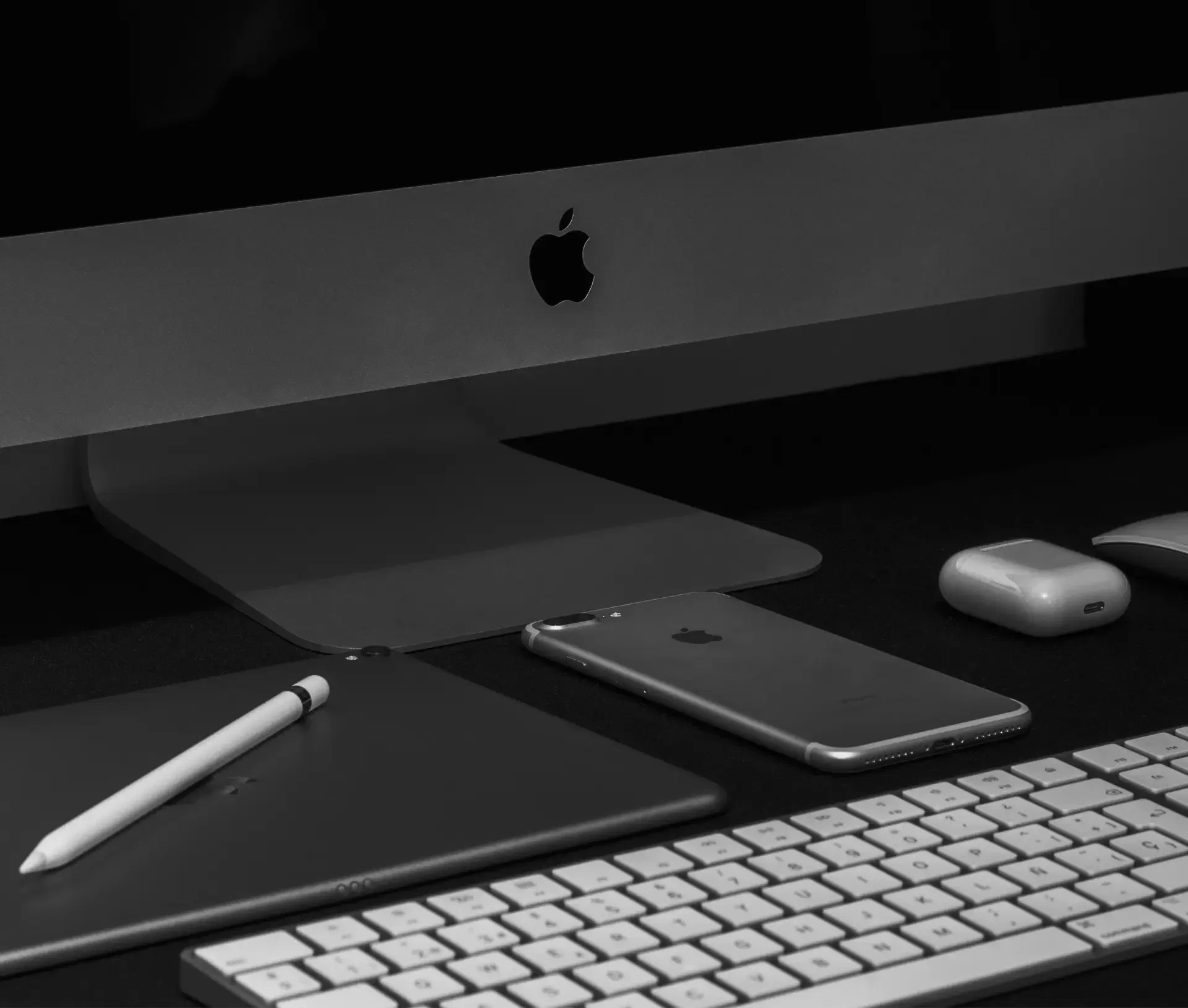A black and white photo showing an Apple iMac, wireless keyboard, iPhone, iPad with Apple Pencil, and AirPods on a desk. Perfect for showcasing Apple Business solutions supported by Jamf Free MDM. The Apple logo is visible on the iMac and iPad.