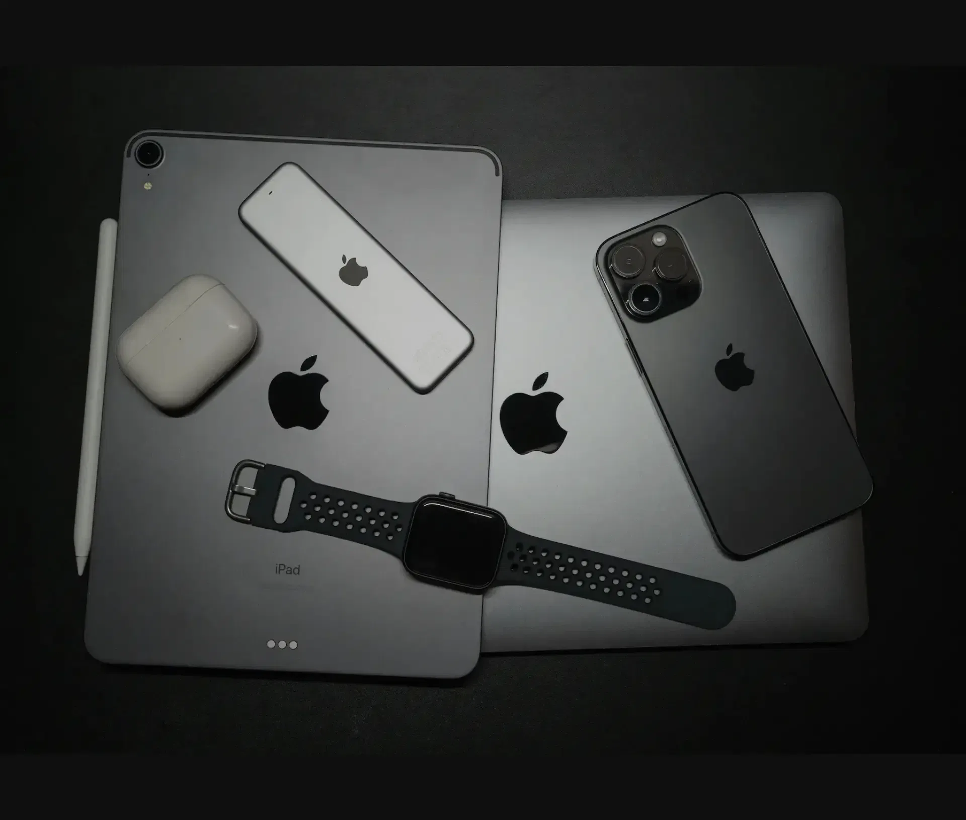 A collection of Apple devices on a dark surface, including an iPad, MacBook, Apple Watch, iPhone, AirPods, Apple Pencil, and an iPod—ideal for creative teams collaborating while staying mindful of cyber security risks.