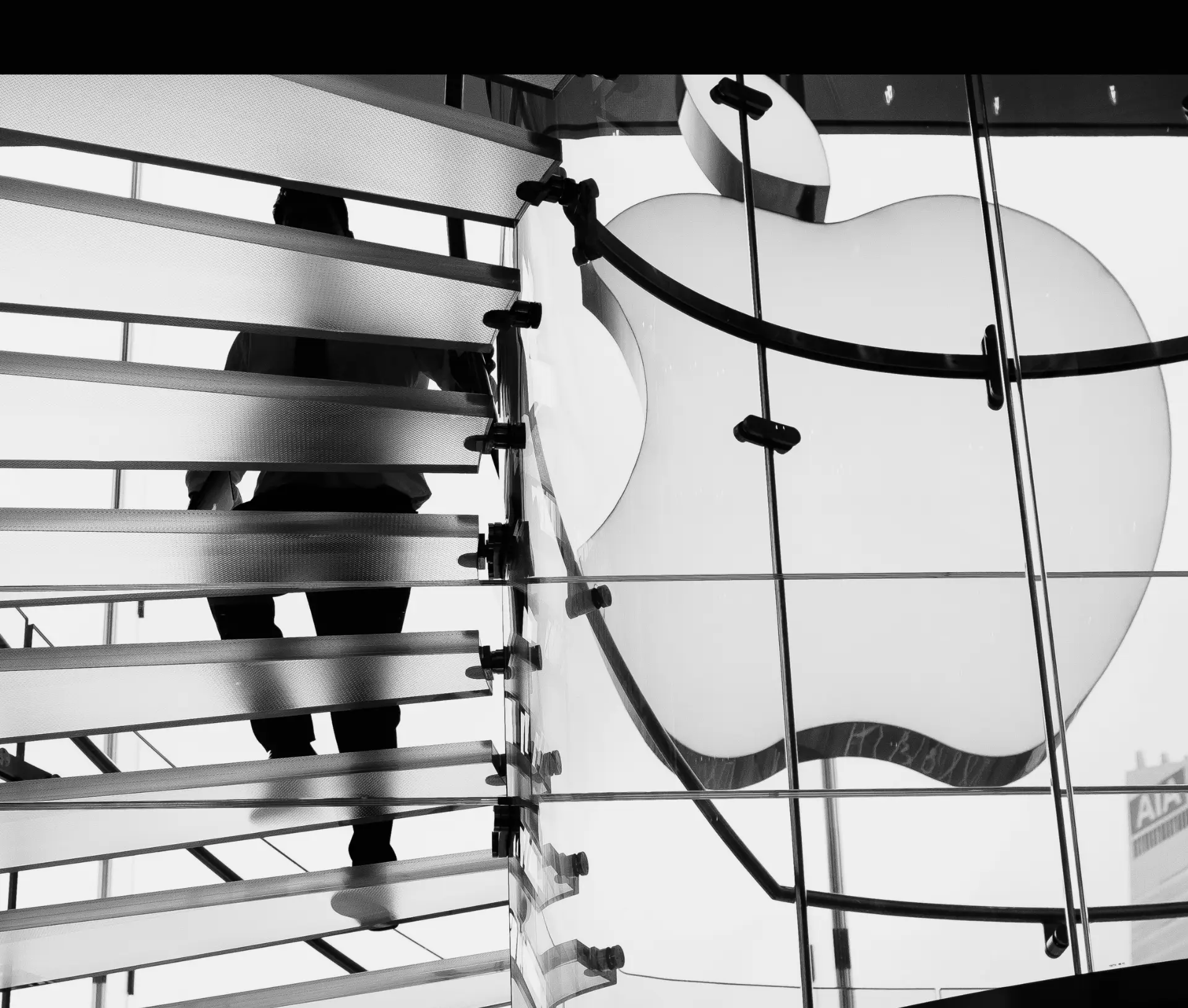 A person walks up a glass staircase beside a large illuminated Apple logo inside an Apple Store, evoking the innovation behind Apple Business and third party MDM solutions. The black and white scene features the figure partially silhouetted.