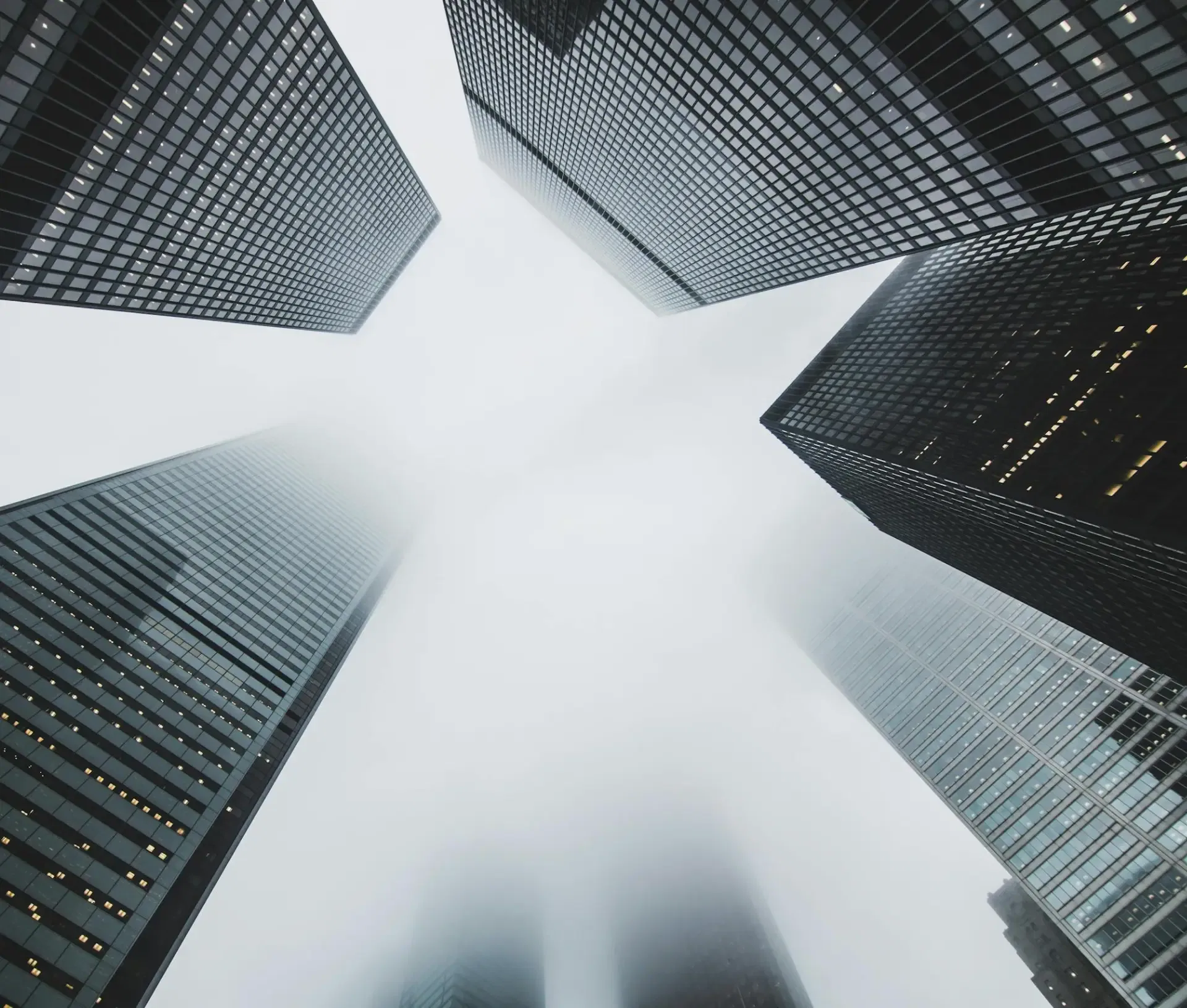 View looking up at tall skyscrapers shrouded in mist, with the tops obscured by fog and a bright, cloudy sky above—much like how an MSP or Apple centric IT partner would evaluate challenges hidden at the heights of modern business environments.