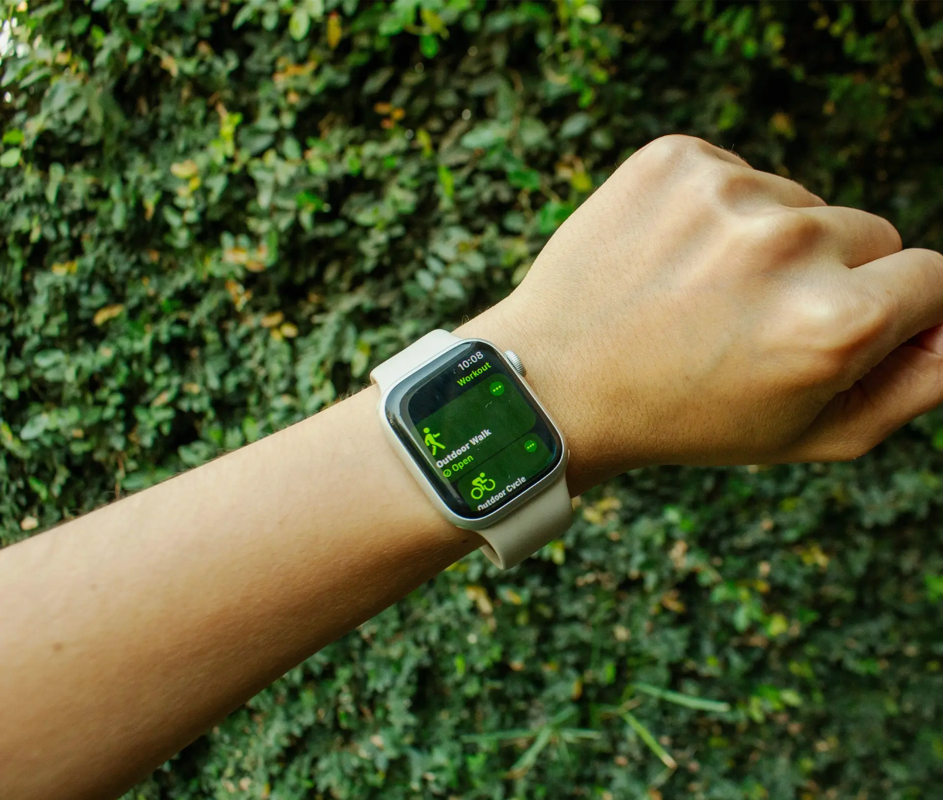 A person’s wrist wearing a smartwatch with a white strap, displaying fitness tracking info for walking and cycling—highlighting built in features from the Apple wellbeing toolkit against a leafy green background.