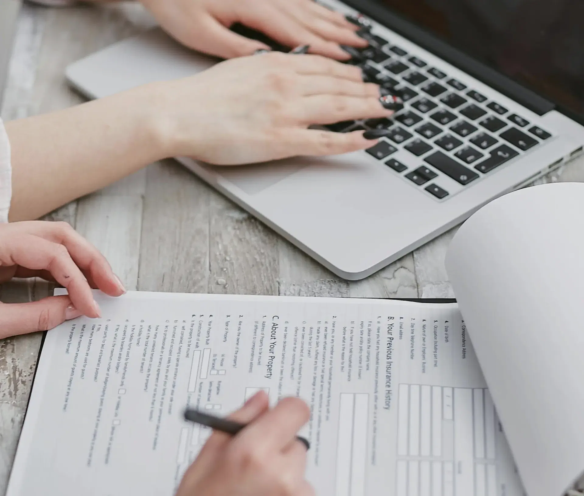 Two people working at a desk: one typing on a macOS laptop, the other filling out a printed compliance form with a pen. The wooden desk has papers spread out next to the Apple device, ideal for regulated industries.