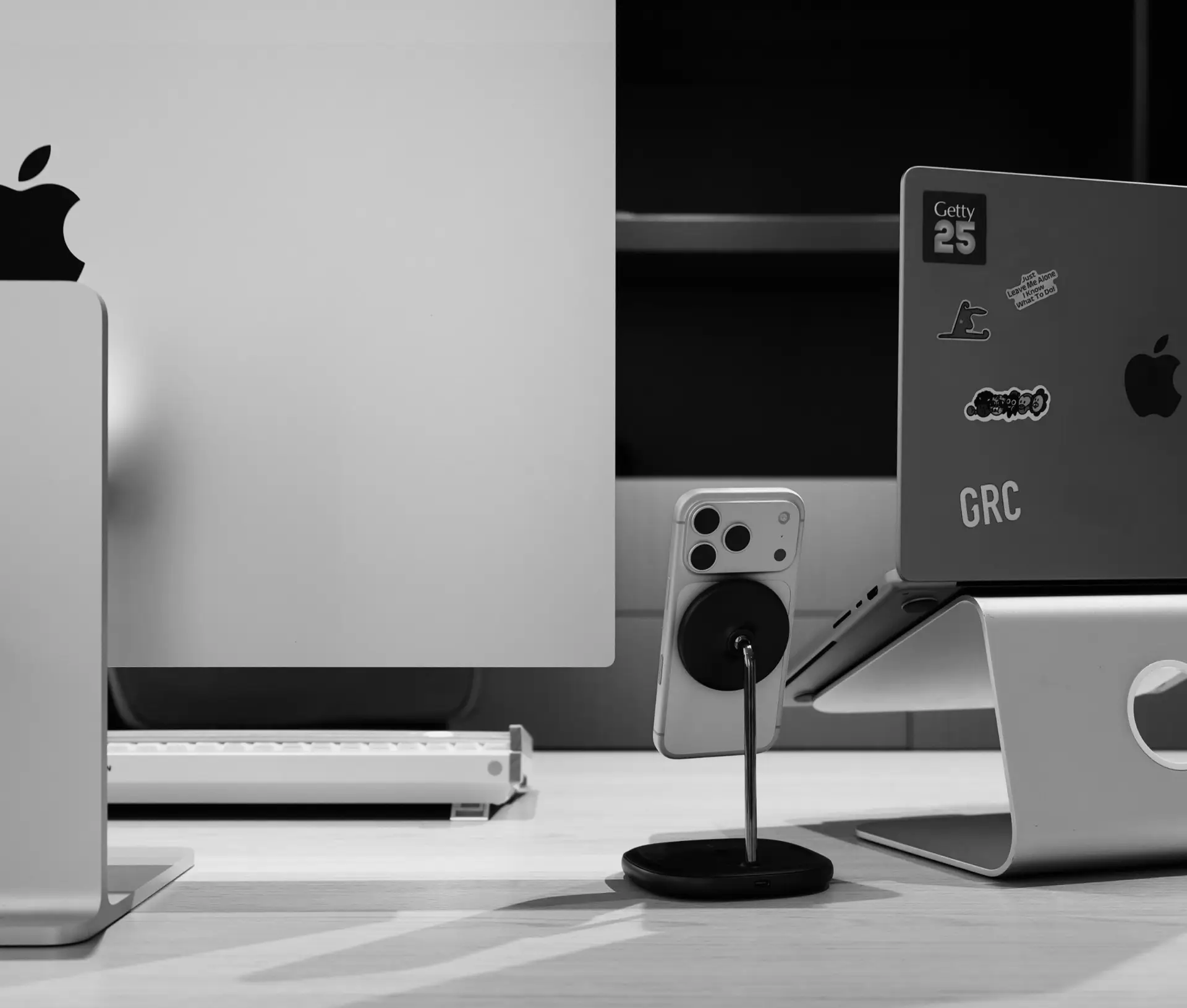 A black and white photo of a modern desk setup highlights an iMac, a MacBook on a stand with stickers, and an iPhone on a wireless charging stand—showcasing Apple Continuity to boost productivity for remote teams. Shelves and part of a keyboard form the backdrop.