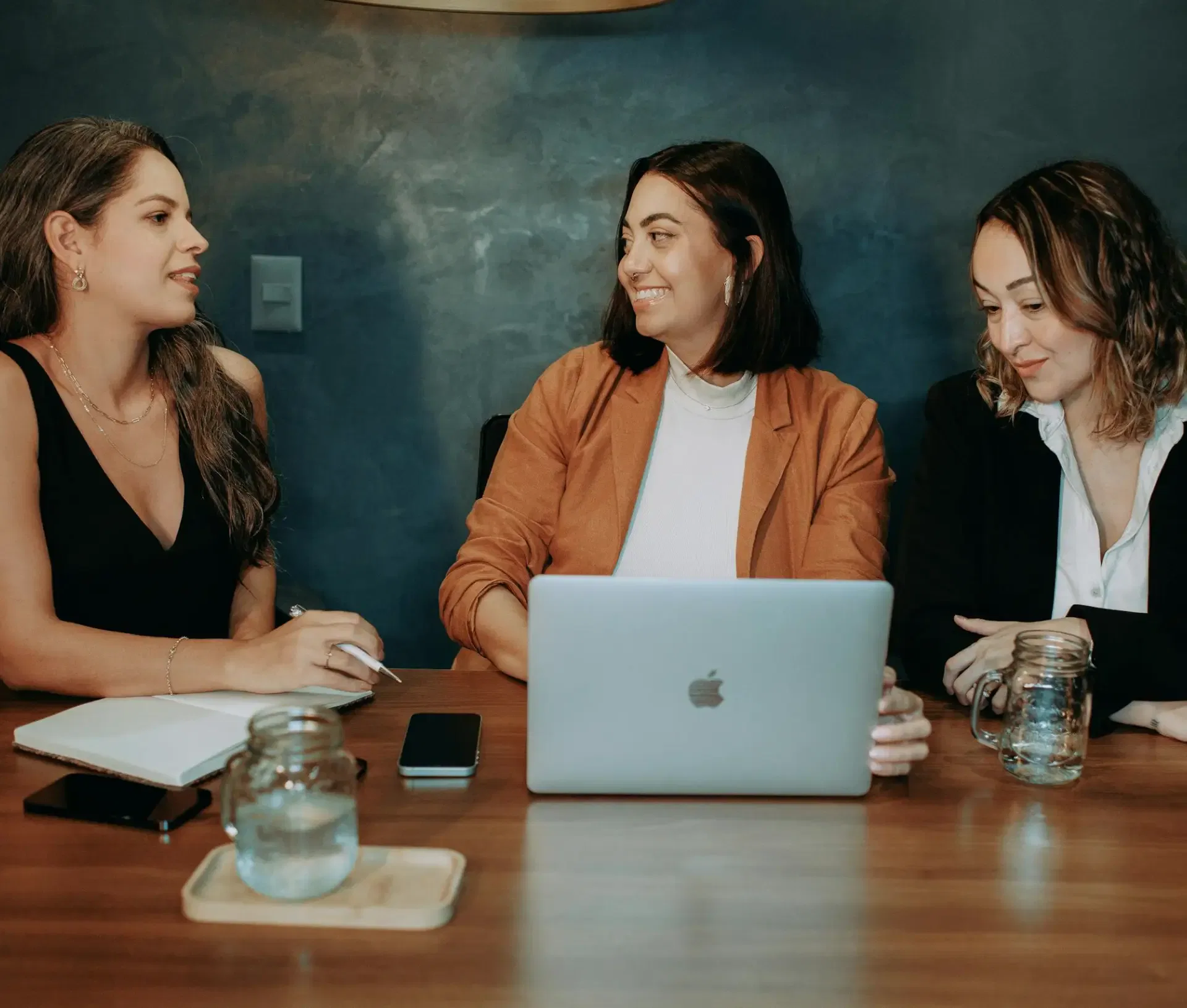 Three women sit at a wooden table with a laptop, notebook, phone, and jars of water. Dressed in business attire, they discuss IT solutions and strategic partnership opportunities in a modern office with a dark blue wall.