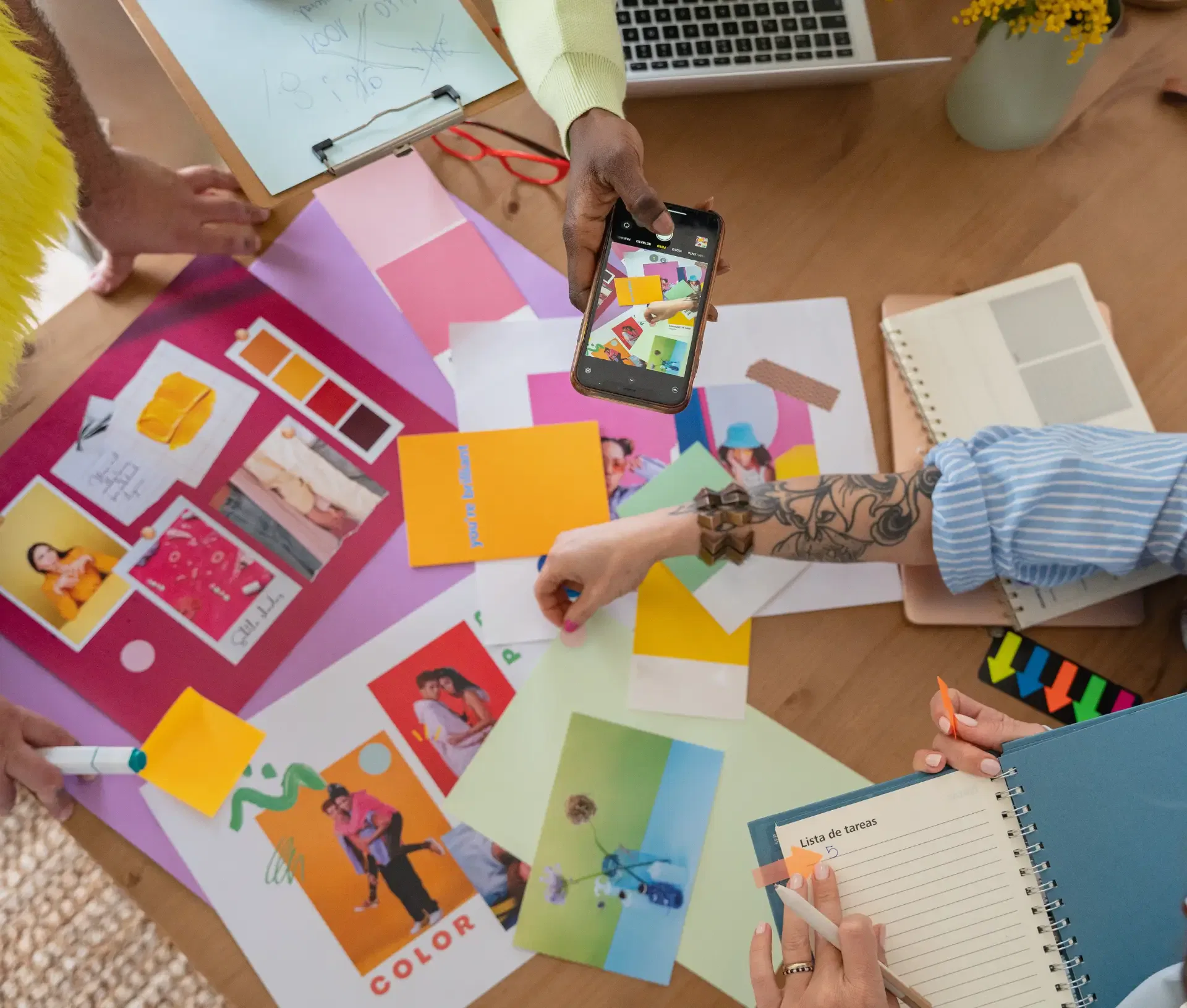 A creative team collaborates at a table filled with colourful design materials, colour swatches, photos, a laptop, and notebooks. One person takes a photo with a mobile as others write and point—mindful of cyber security risks when sharing digital content.