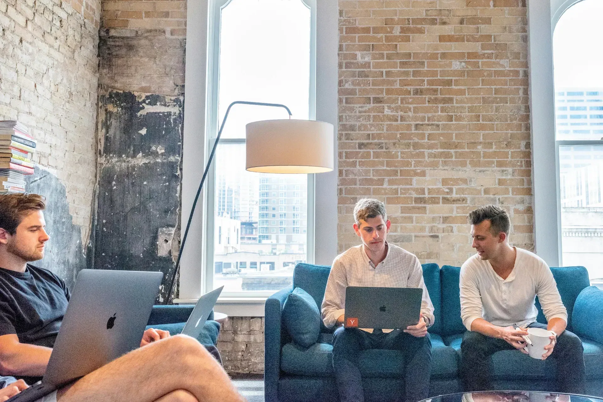 Four people are seated on sofas in a brick walled room with laptops and a cup, engaged in a casual meeting.