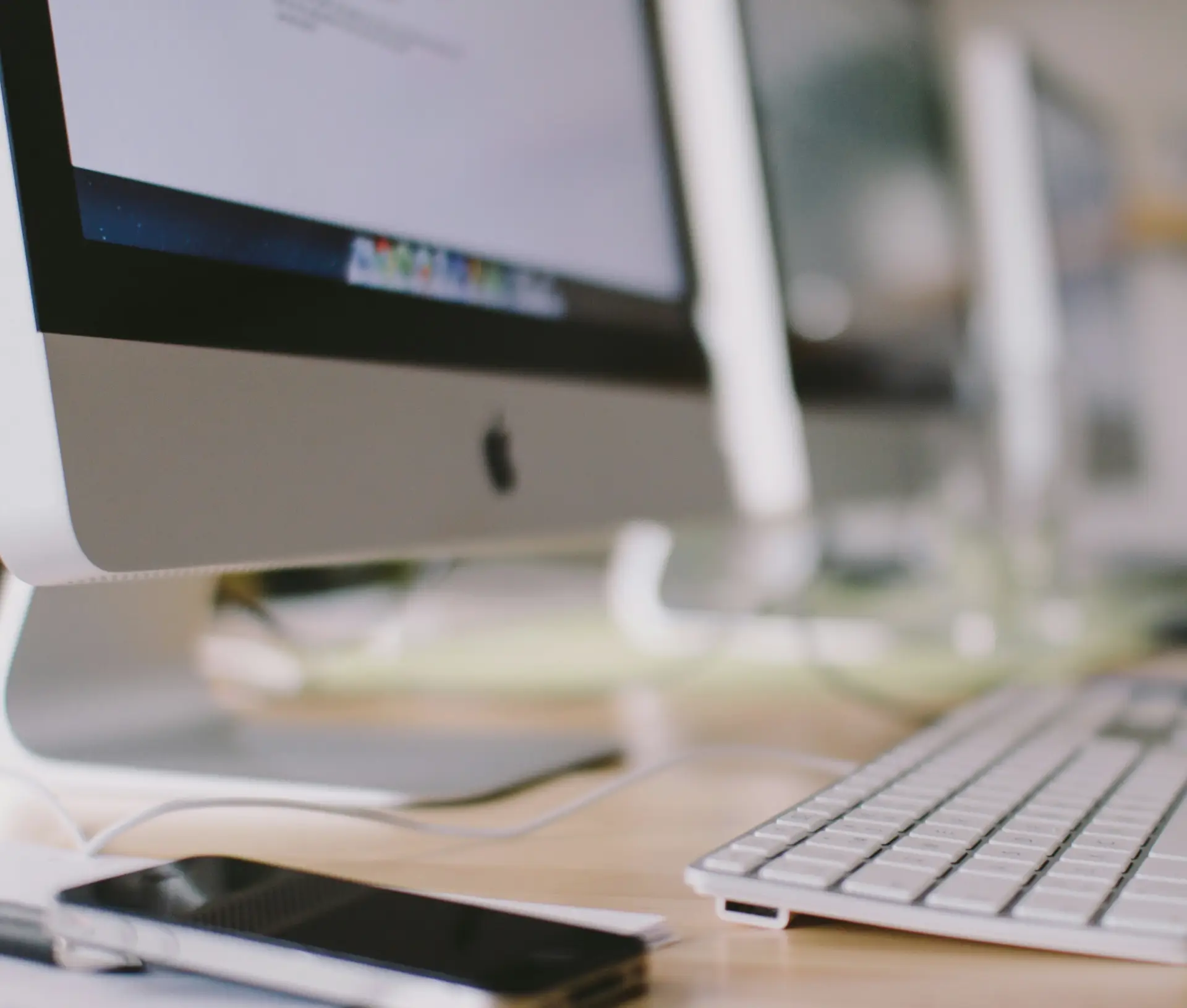A close up of an Apple iMac computer with a wireless keyboard and a smartphone on a desk in a well lit, All Mac Office. Several computer monitors are lined up in the background, reflecting the team’s commitment to cyber security.