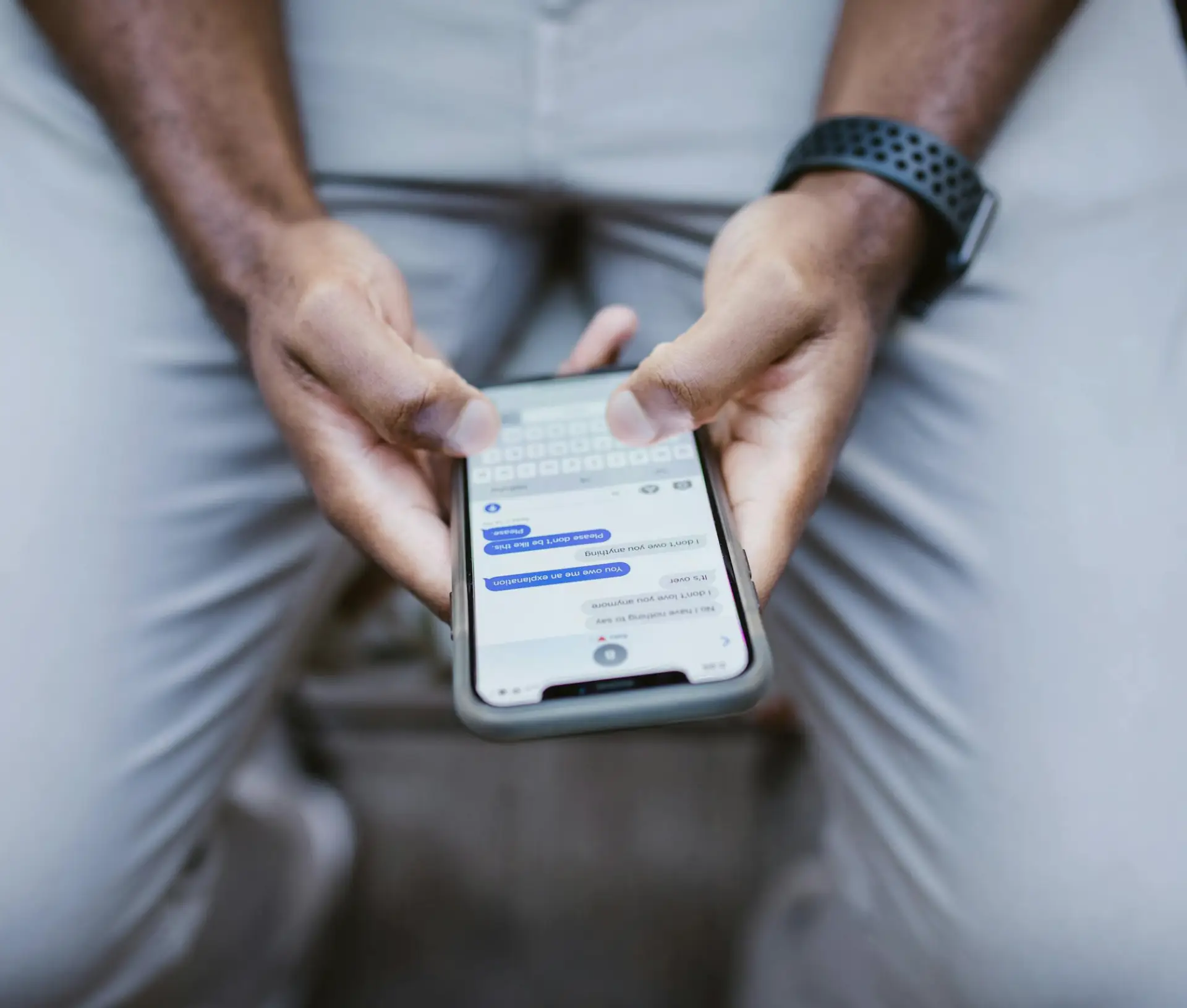 A person wearing a black watch and light trousers is holding a mobile phone with both hands, typing a message in an encrypted RCS messaging app. The phone screen shows a secure text conversation.
