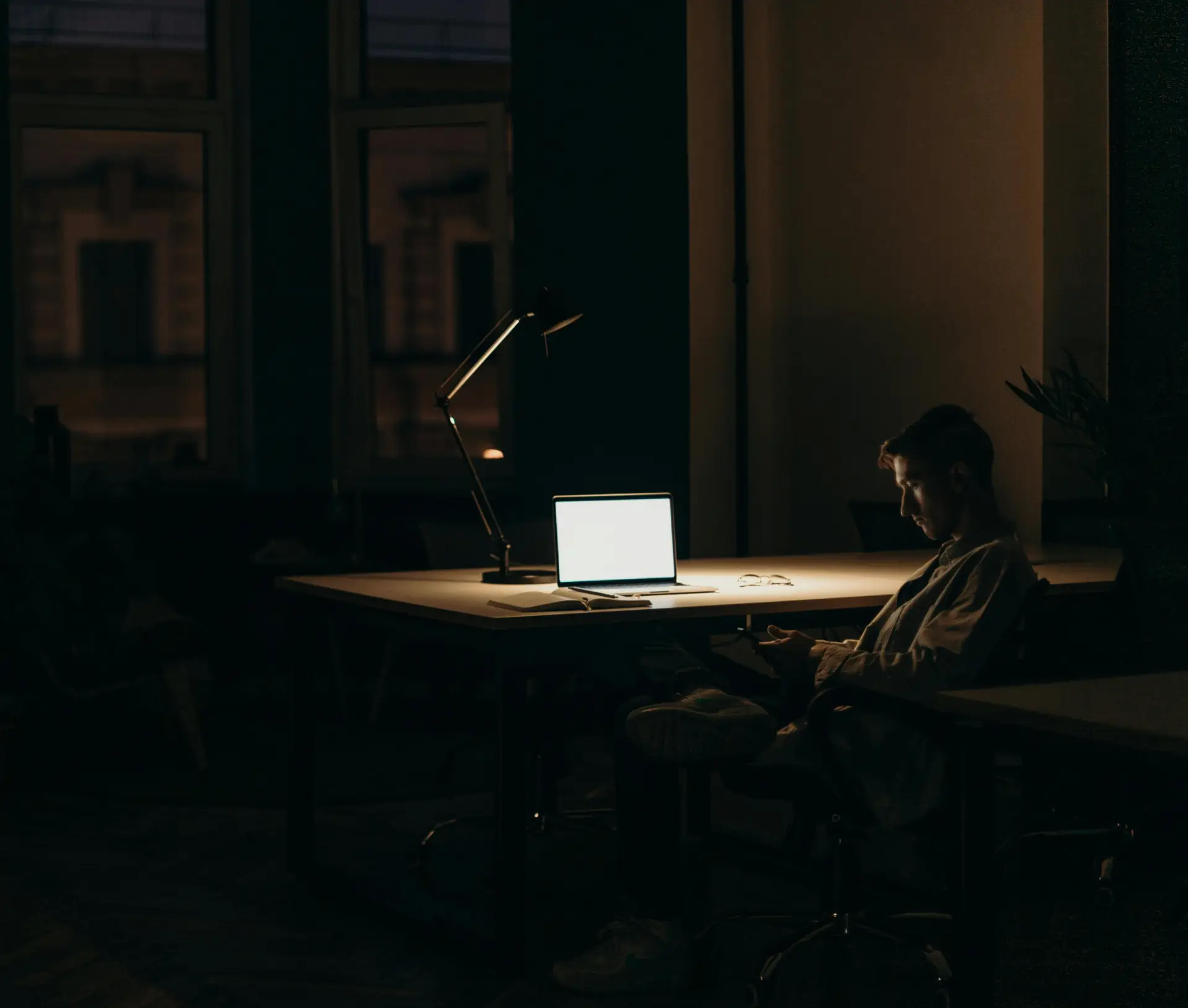 A person sits alone in a dark office at night, illuminated by their laptop running macOS Focus Modes. Glancing at a mobile, they seek ways to reduce burnout—a quiet moment amidst remote teams and glowing city lights beyond the windows.