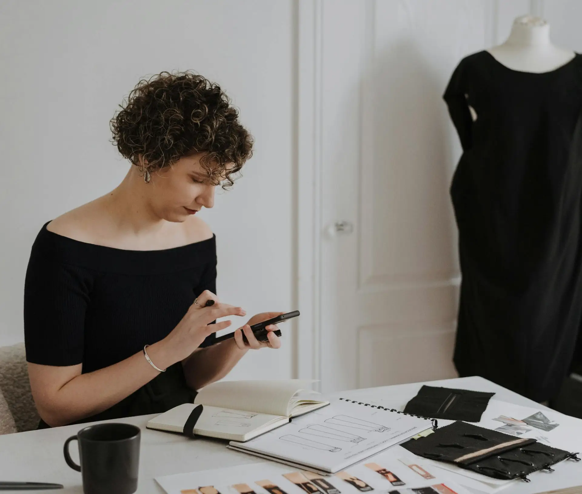 A person with curly hair sits at a desk using a mobile phone, surrounded by design sketches, fabric samples, and a notebook—focused on securing creative file breaches. A black dress on a mannequin stands in the background.