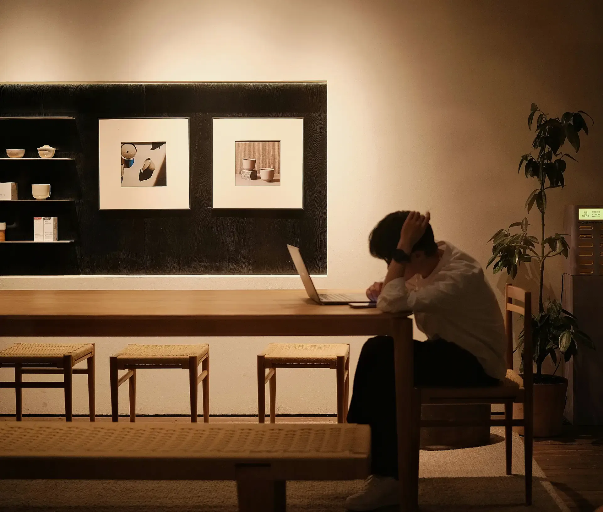 A person sits alone at a wooden table, leaning over a laptop with their hand on their head. The dimly lit room has minimalist decor with framed art, shelves, and a potted plant—reflecting the challenges of business technology and IT satisfaction.