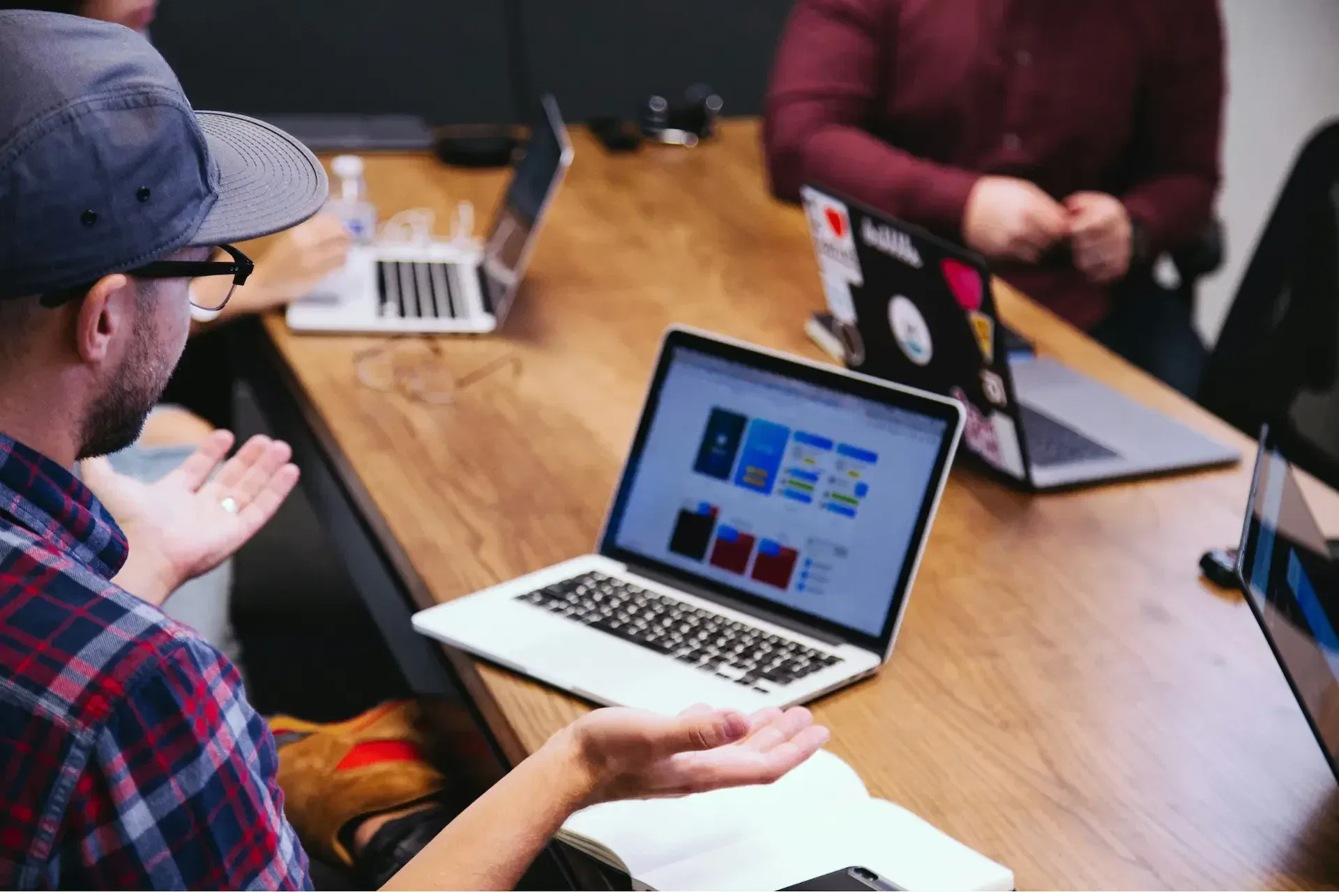 Three people sit at a wooden table with open laptops, engaged in discussion; one person gestures with hands while others focus on their screens.
