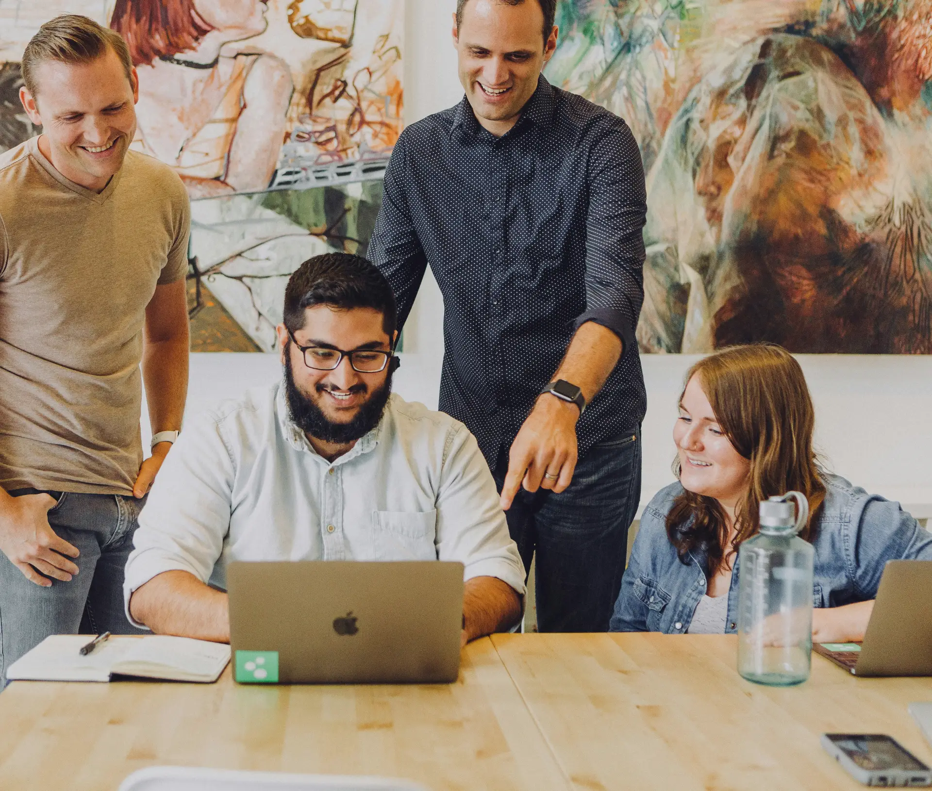 Four people gather around a table with laptops, collaborating on Jamf solutions for UK businesses. One person is seated, typing, whilst another points at the screen. The atmosphere is friendly and casual, with colourful artwork in the background.
