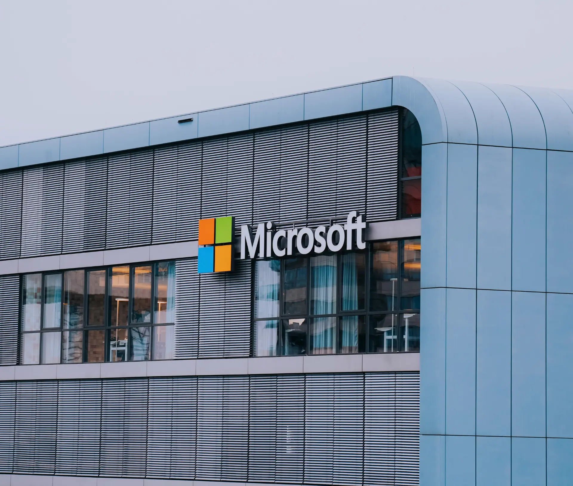 The exterior of a modern building featuring the Microsoft logo and name, with large windows and a sleek, metallic façade under an overcast sky—highlighting enhanced security measures like MFA.