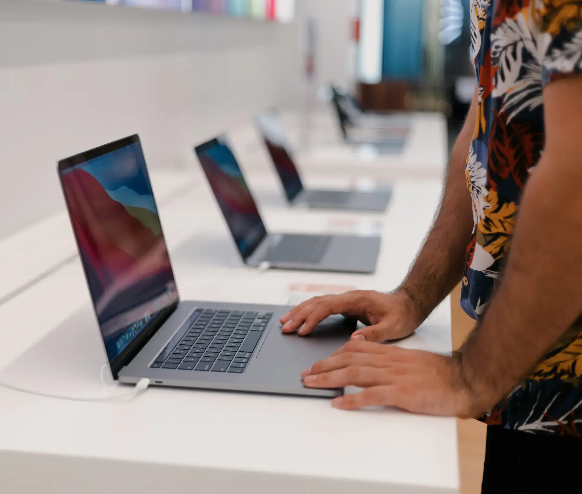 A person in a floral shirt uses a laptop on display at a shop, exploring options such as Lease vs. Buy through Apple Financial Services, with several other laptops lined up on the white counter—an approach to scaling in 2026.