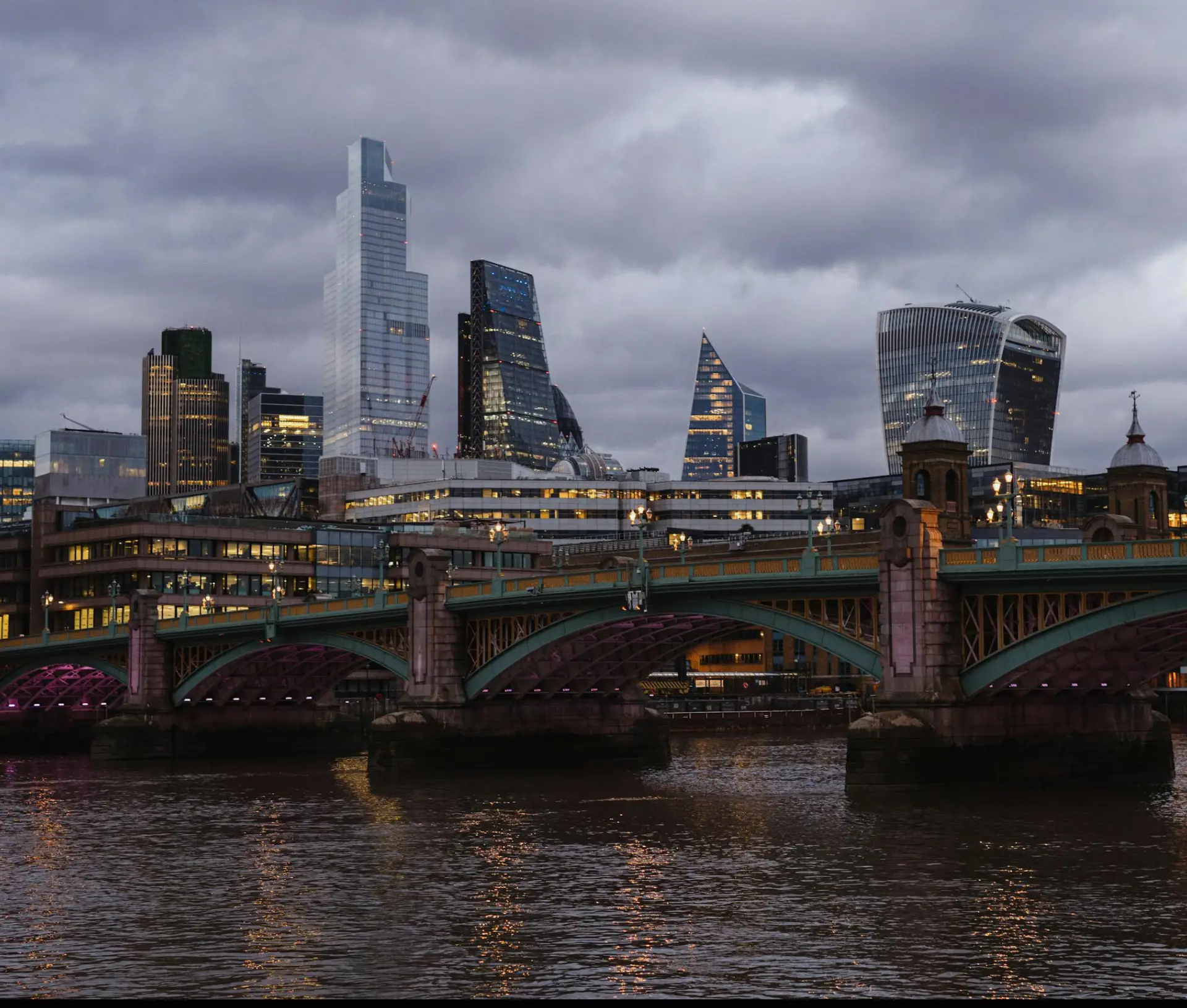 A cloudy evening view of London’s city skyline with modern skyscrapers behind Southwark Bridge, which crosses the Thames and is lit with purple lights—perfect for deep work or to help reduce burnout using macOS Focus Modes.