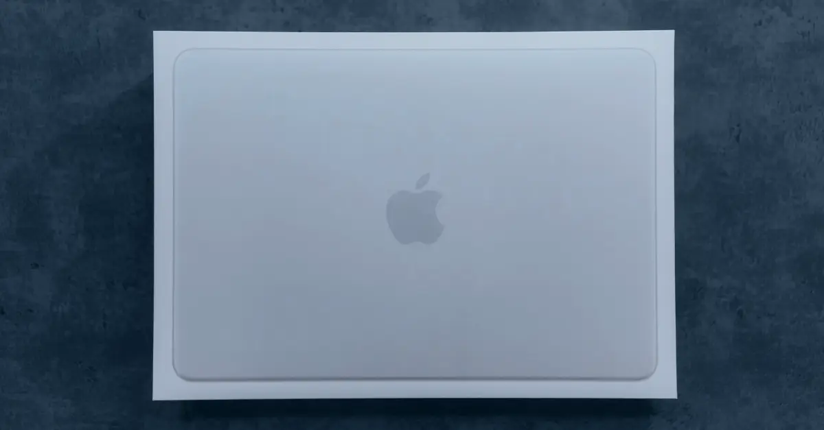 A white Apple product box with a subtle Apple logo in the centre sits on a dark grey surface, viewed from above—perfect for your next MacBook Neo business review.