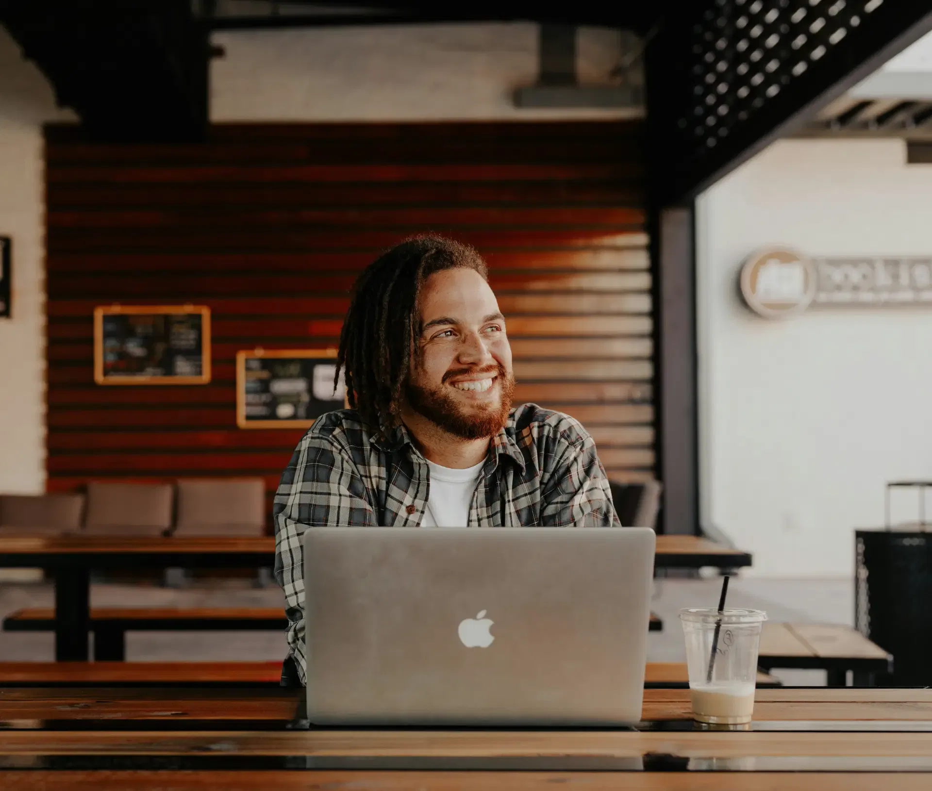 A man with dreadlocks and a beard smiles whilst sitting at a table with a laptop and drink in a modern, outdoor café setting, reflecting on IT decisions that promote workplace wellbeing.