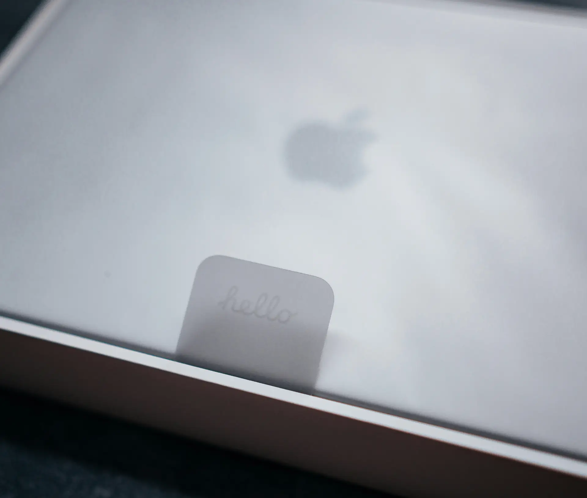 A silver Apple laptop inside its box with a translucent sheet on top and a small tab labelled 
