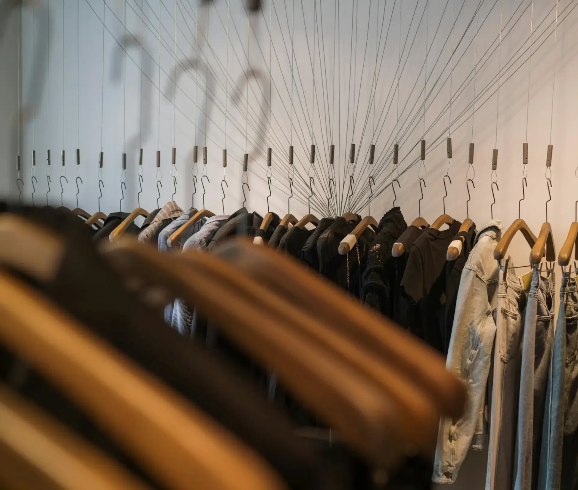 A clothes rail displays various garments on wooden hangers, arranged in a row. Above, empty hangers are suspended by lines from the ceiling against a white wall, creating a modern retail shop scene that highlights innovative Retail Technology.