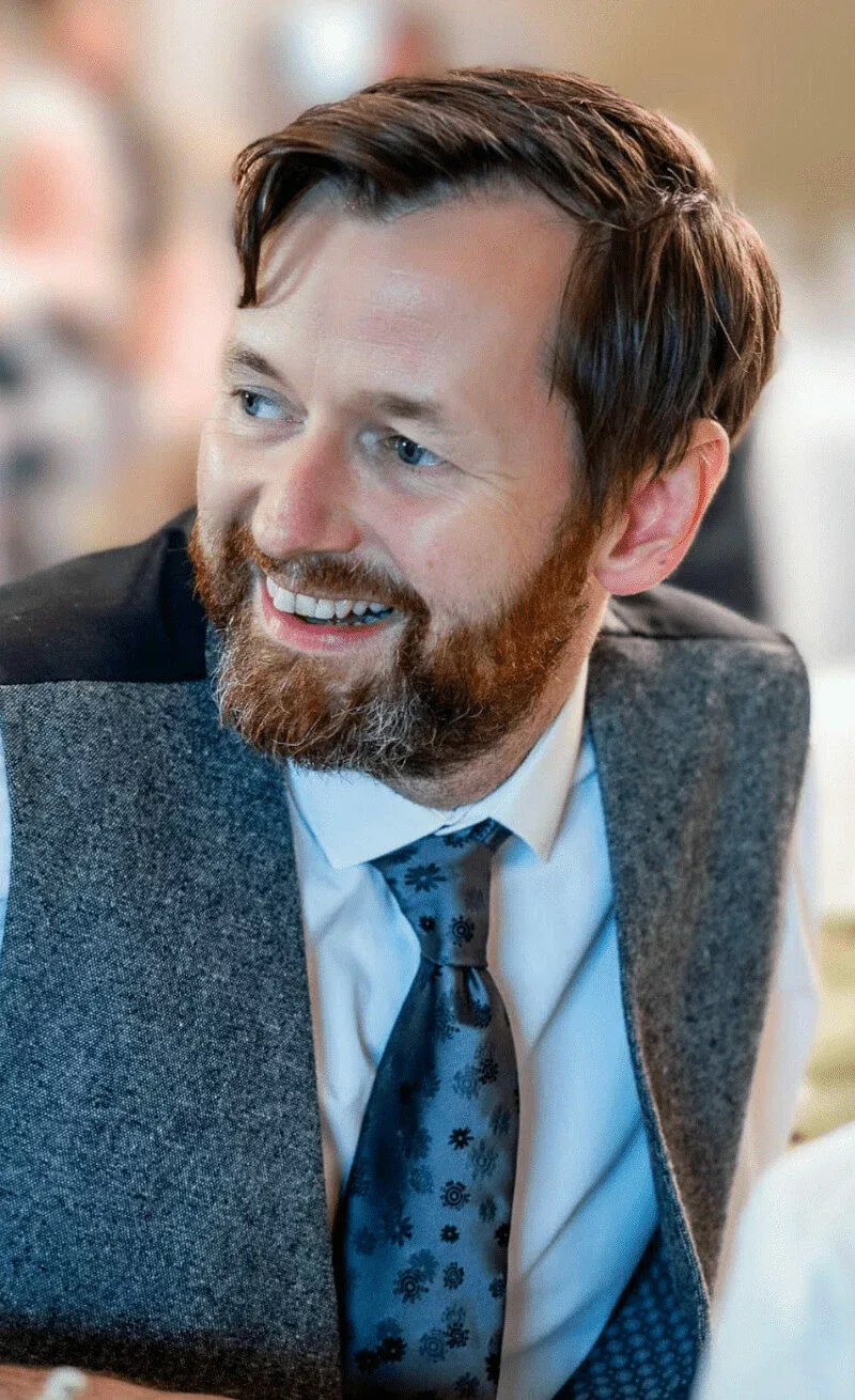 Bearded man in a gray vest and blue patterned tie smiling, looking to the side.