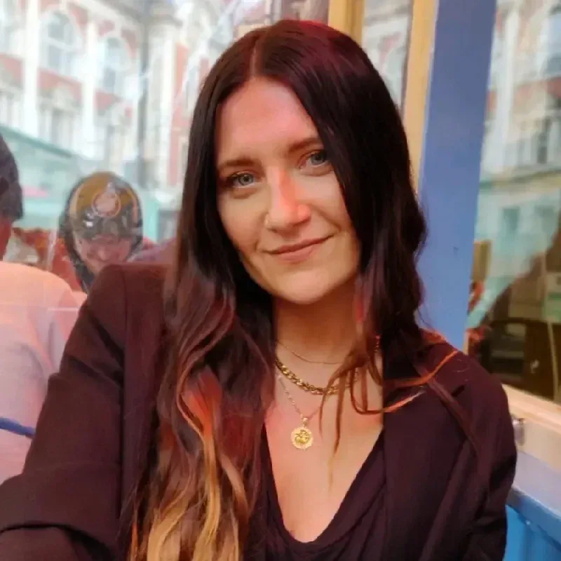 Woman with long dark hair and layered necklaces sits at an outdoor cafe table, with buildings visible in the background.