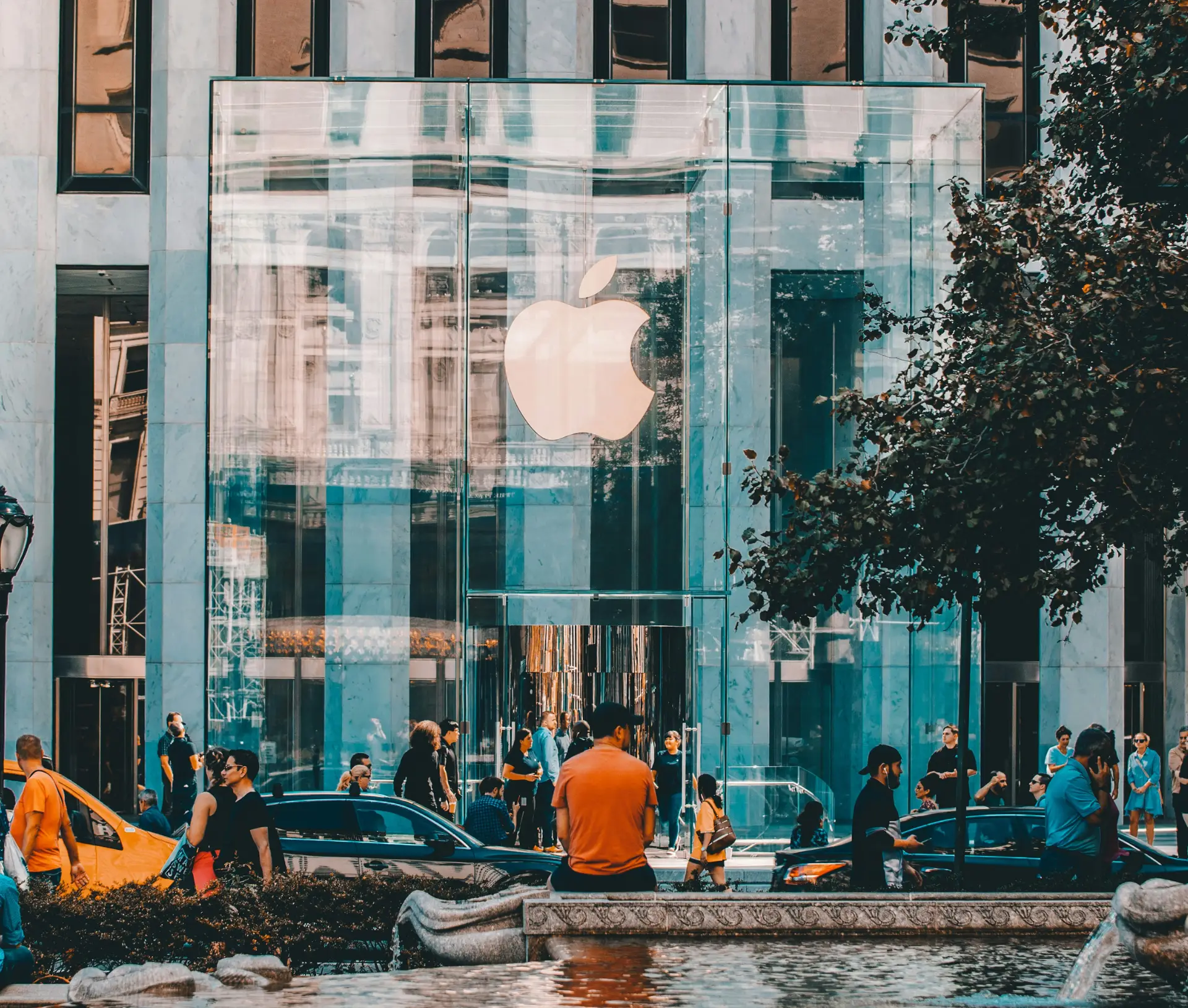 People walk and gather outside a large glass fronted Apple Store with the white Apple logo prominently displayed above the entrance, showcasing the vitality of Apple businesses as cars and trees line the bustling city street.