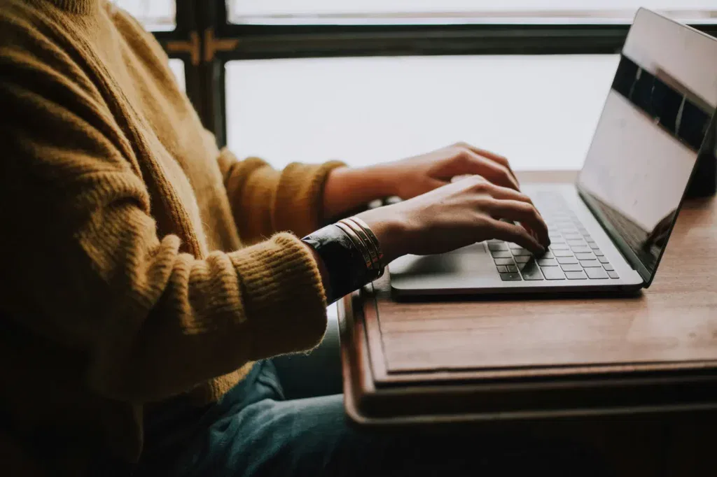 Person wearing a yellow sweater typing on a laptop at a wooden table near a window.