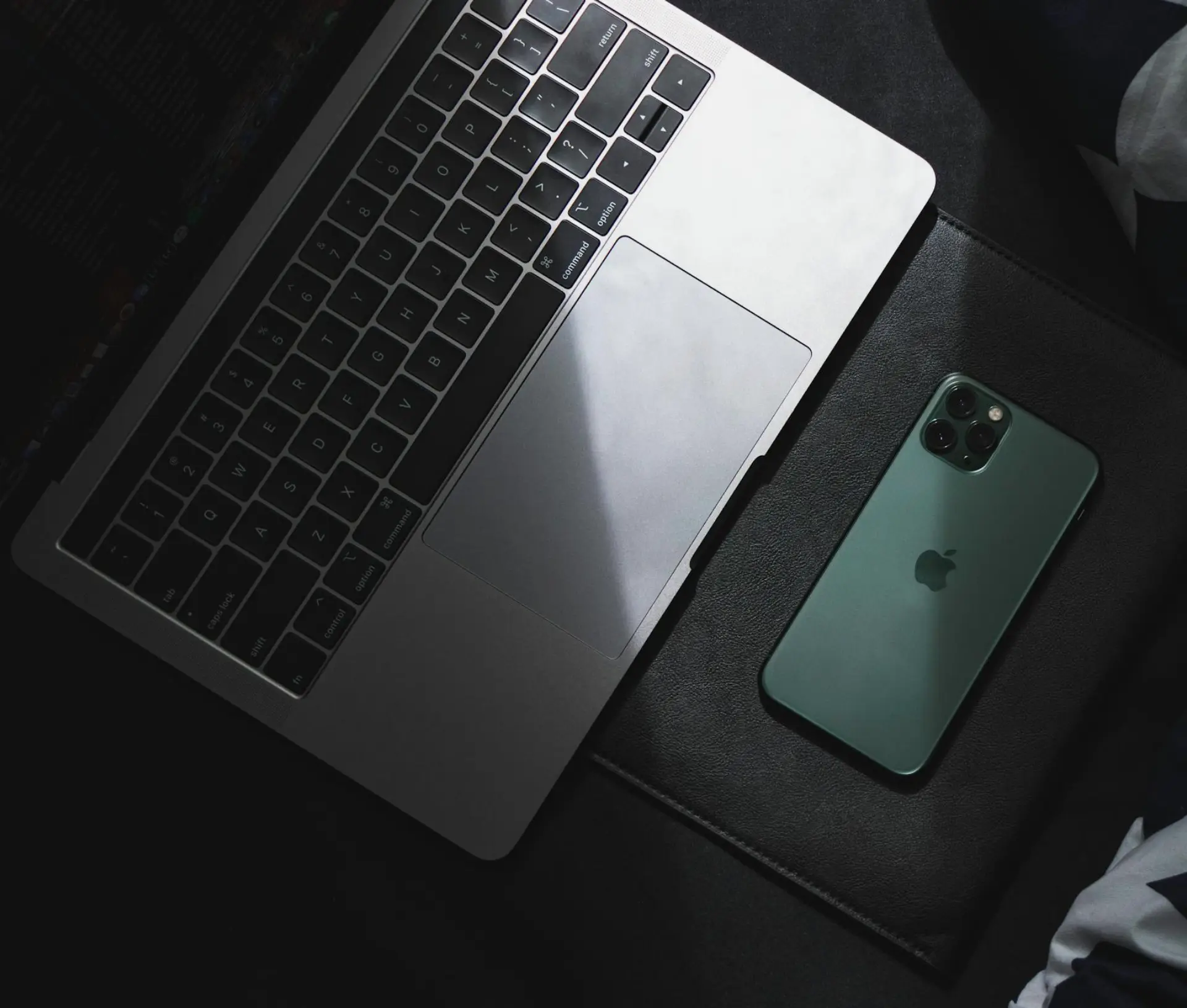 A silver laptop and a green iPhone, ready for the latest iOS update, are placed side by side on a dark surface, with light partially illuminating the laptop’s keyboard and trackpad.