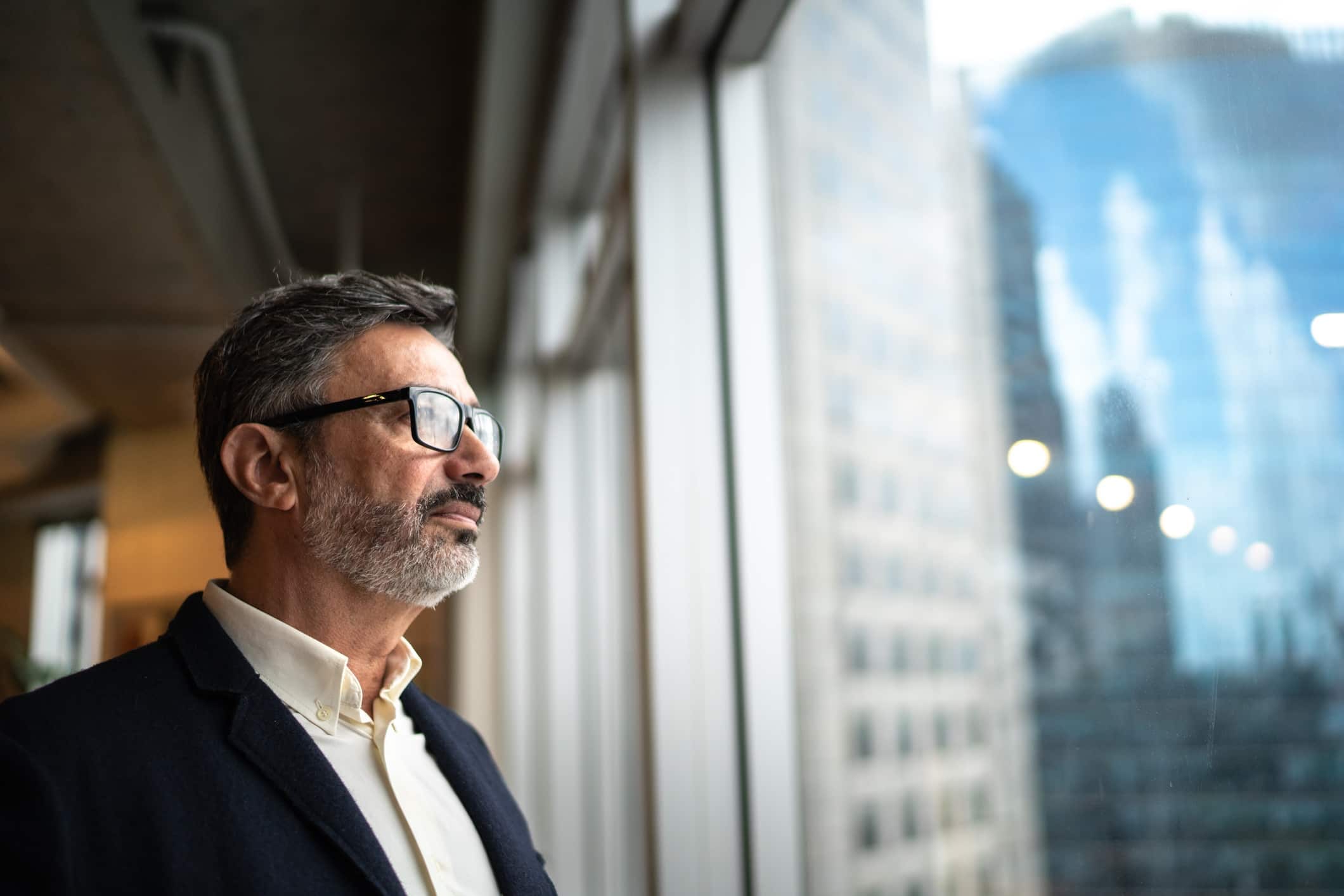 Man wearing glasses and a suit looks out of a large window in an office building.