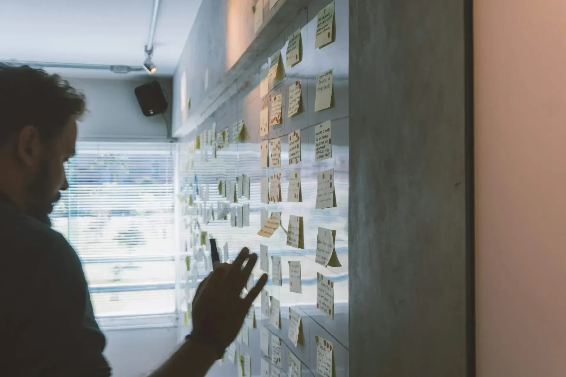 A person stands in front of a wall covered with numerous sticky notes, some written on, in a brightly lit room with large windows, preparing for quarterly business reviews led by the virtual cio.