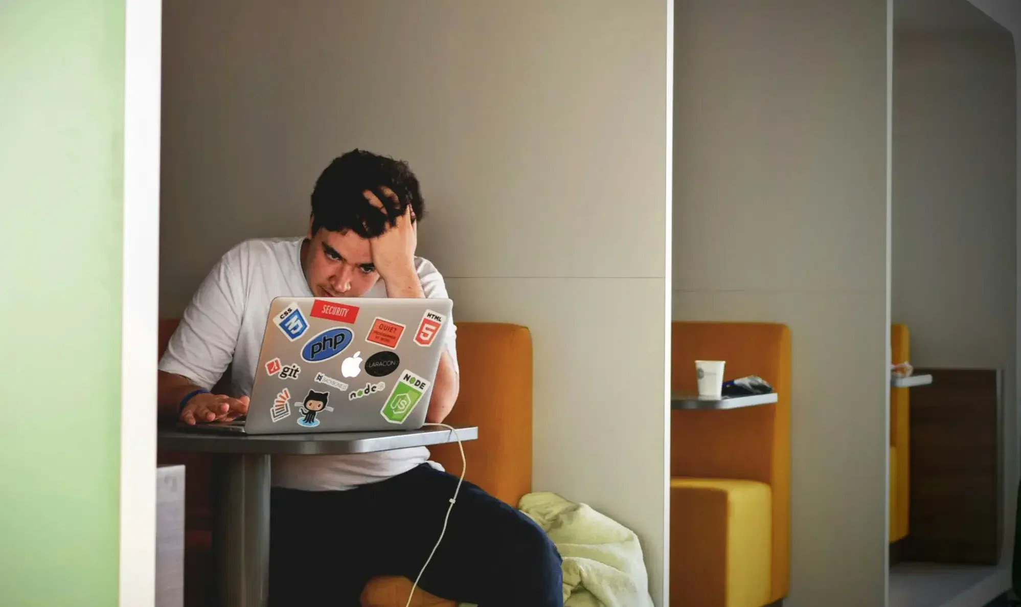 A person sits at a small table in a booth with an open laptop covered in tech stickers, resting their head in one hand and showing signs of Tech Stress Test. A drinks cup is visible on a nearby table.